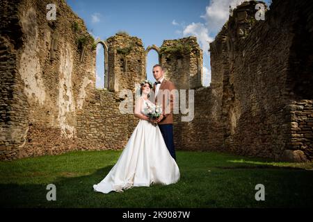 A lovely couple in a suit and a wedding dress poses for camera next to old stone walls of the castle Stock Photo