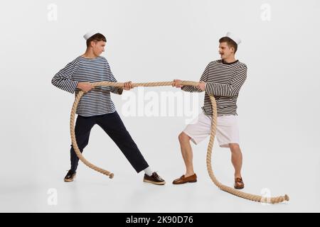 Portrait of two young boys, sailor, seamen in striped shirts pulling rope isolated over white background. Competition Stock Photo
