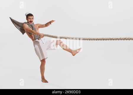 Portrait of young cheerful man, sailor in striped shirt on shoulders pulling rope isolated over white background Stock Photo