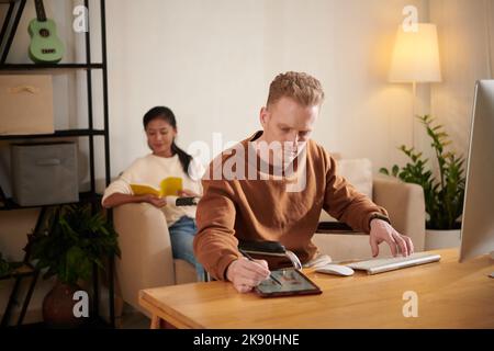 Talented creative young man in wheelchair drawing fashion sketches, his wife reading in background Stock Photo