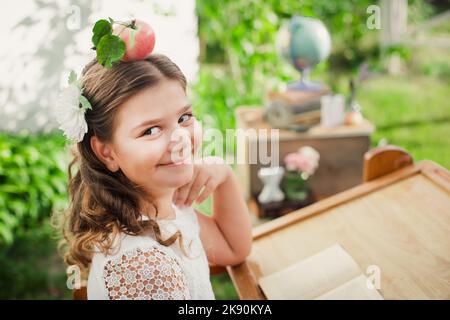 Happy smart schoolgirl in school unifrom hold red apple on head. student sits at desk against background of green grass and globe. Stock Photo