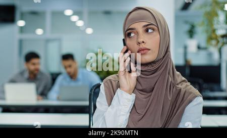 Serious focused Arabic female millennial manager in hijab talking on phone, consulting, making business call at work, young female worker or customer Stock Photo