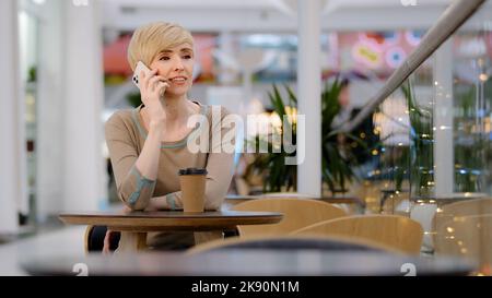 Caucasian mature adult middle aged 40s woman business lady sitting at table in cafe talking on mobile phone smartphone answering call online order Stock Photo