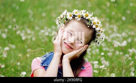 On a chamomile lawn, a sweet girl in a wreath of daisies, smiling, pressing her hands to her cheeks. High quality photo Stock Photo