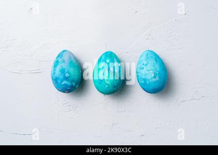 Three blue and turquoise hand-painted Easter eggs lined up on a white textured background with copy space. Stock Photo