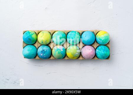 Hand-painted colorful Easter eggs in a cardboard carton on a white textured background, top view. Stock Photo