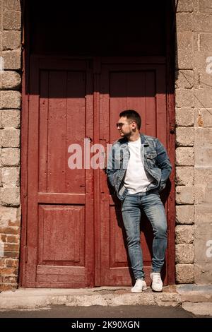 Handsome man with a beard in a denim jacket and denim pants stands near red vintage doors. Grown guy in denim style Stock Photo