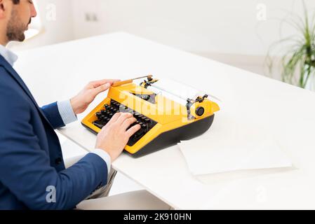 Close-up of a man typing on retro yellow typewriter while sitting at desk. Blank paper. Stock Photo