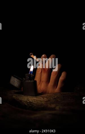 A hand holding a cigarette with a lit lighter at night in a vertical shot Stock Photo