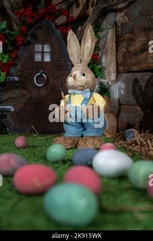 A vertical shot of a cute straw Easter bunny wearing blue trousers with colorful eggs in the foreground Stock Photo