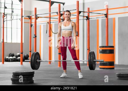Full length of young strong female athlete with dark hair in sportswear smiling happily and looking at camera while standing near heavy barbell in mod Stock Photo