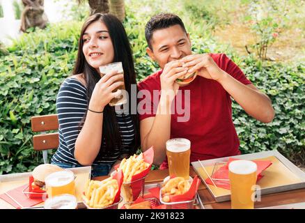 Couple eating tasty burger and drinking alcoholic beer while sitting on terrace of outdoor cafe with green plants in city Stock Photo