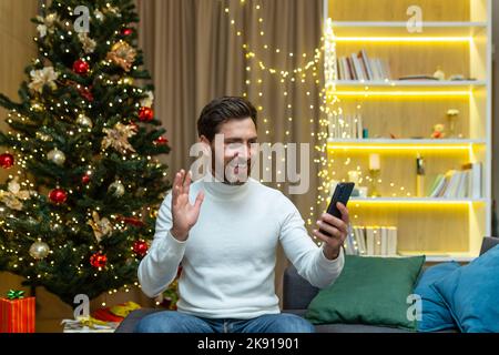 Businessman at home for Christmas and New Year holidays sitting on sofa in living room near Christmas tree talking on video call, man having fun talking with friends holding phone Stock Photo