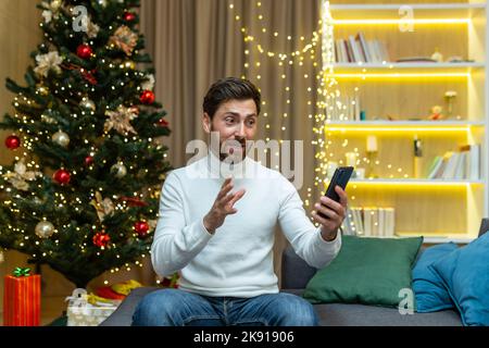 Businessman at home for Christmas and New Year holidays sitting on sofa in living room near Christmas tree talking on video call, man having fun talking with friends holding phone Stock Photo