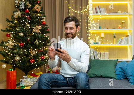 Cheerful man at home uses the phone, celebrates Christmas and New Year sitting on the sofa near the Christmas tree, types messages and browses online on the Internet. Stock Photo