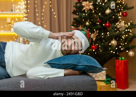 A man is tired for Christmas, in a New Year's hat, he is sleeping by the Christmas tree on the sofa at home in the living room after celebrating Christmas, he has a bad headache from a hangover. Stock Photo