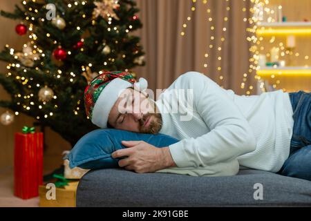 A man is tired for Christmas, wearing a New Year's hat, sleeping near the Christmas tree, sitting on the sofa at home in the living room after celebrating Christmas. Stock Photo