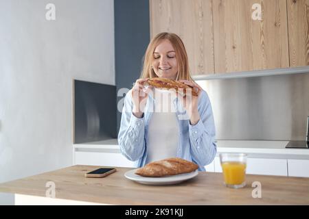 Close-up portrait of happy healthy woman eats freshly baked baguette with prosciutto for breakfast in morning in light white interior style kitchen Stock Photo