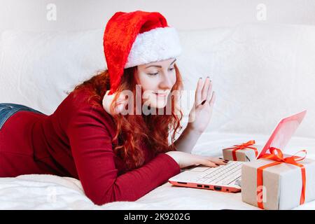 Young woman in Santa hat is lying on couch with laptop. Cheerful lady uses video chat for online greetings for Christmas and Happy New Year. Stock Photo