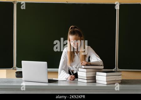 Education in school. Schoolgirl with laptop and books making homework. Girl at school board study and writing notes in notebook Stock Photo