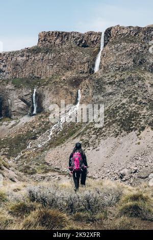 Rear view of female backpacker hiking through a rocky path to a big waterfall. Vertical shot Stock Photo