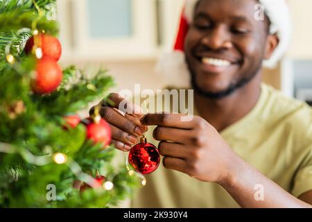 afro american man in Santa hft decorate the christmas tree with a red ball at home Stock Photo