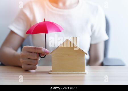 Businesswoman hand holding red Umbrella cover wooden Home model. real estate, insurance and property concepts Stock Photo