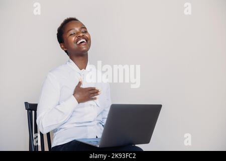 African-american overjoyed happy woman holding, using, working on laptop pc computer sitting on chair isolated over white wall. White wall background Stock Photo