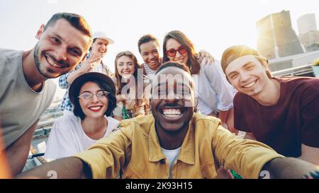 Point of view shot of cheerful young people multiethnic group taking selfie and holding camera, men and women are looking at camera, smiling and posing with drinks at rooftop party. Stock Photo