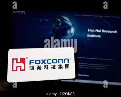 Person holding cellphone with logo of Hon Hai Precision Industry Co. Ltd. (Foxconn) on screen in front of webpage. Focus on phone display. Stock Photo