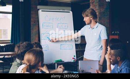 Creative manager is using whiteboard at business meeting in office pointing at chart and talking while group of people is listening and learning new information. Stock Photo