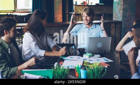 Handsome young man director is talking to his employees and gesturing sitting together at desk in modern office. Laptop, documents with charts and graphs are visible. Stock Photo