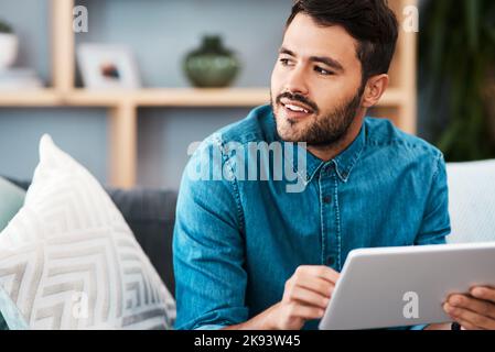What could he be thinking I wonder. a handsome young man looking thoughtful while using a digital tablet in his living room at home. Stock Photo