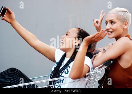 You havent had fun until youve taken some selfies. two cheerful young girlfriends taking a selfie while playing around with a shopping cart outdoors. Stock Photo