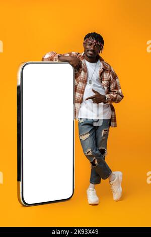 Smiling african man standing near huge smartphone with blank screen on yellow studio background Stock Photo