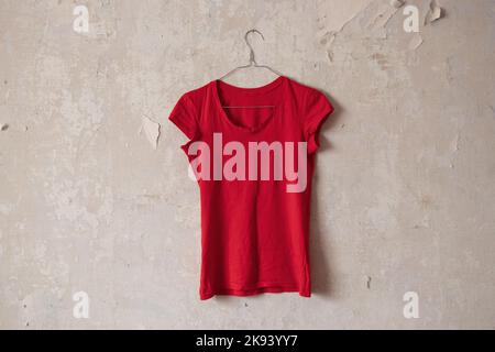 Red womens t-shirt hanging on a hanger on an old dirty white wall at home Stock Photo