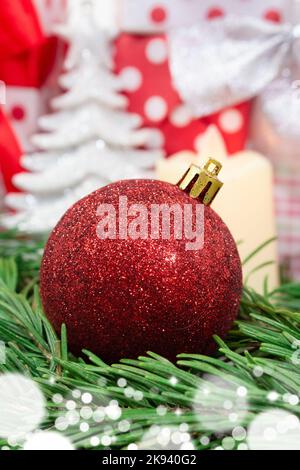 Red shiny ball on fir branches with bokeh, gifts from behind, Christmas tree. Vertical. New Year Stock Photo