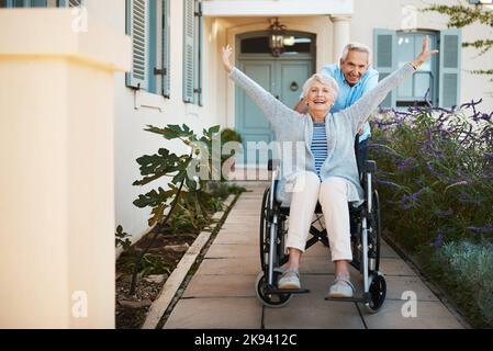 Life is an adventure, enjoy it. Full length shot of a cheerful senior man pushing his wife in a wheelchair in their backyard at home. Stock Photo