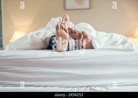 Lets stay in bed for the rest of day. an unrecognizable couples feet poking out under the sheets while lying in bed together at home. Stock Photo