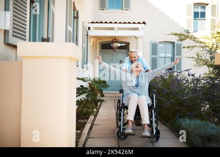 Were still living a life full of thrill. Full length shot of a cheerful senior man pushing his wife in a wheelchair in their backyard at home. Stock Photo