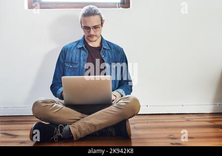 He works wherever theres wifi. Full length shot of a handsome young businessman working on his laptop while sitting on the floor in the office. Stock Photo