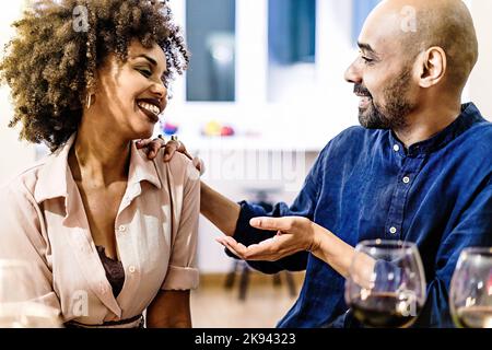 Happy multi-ethnic adult couple flirting sitting on a table at home having fun talking together dining and drinking wine - people and lifestyle concep Stock Photo
