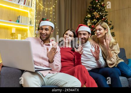 diverse friends celebrating New Year and Christmas together home, Christmas party with guests, men and women smiling and happy together talking on video call greeting friends remotely, using laptop. Stock Photo