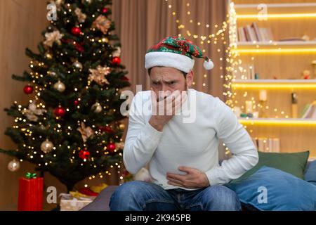 Sick man at home for Christmas, sitting on the sofa in a New Year's hat has severe nausea and stomach pain. Stock Photo