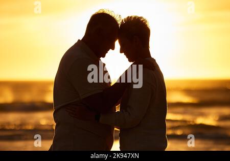 Couple, elderly and silhouette at beach with hug in sunset, evening or dusk by water, waves or horizon together. Senior, man and woman by ocean, sea Stock Photo
