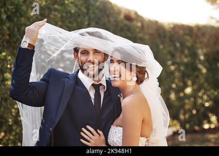 Day one of our happily ever after. an affectionate young newlywed couple smiling while covering themselves with a veil on their wedding day. Stock Photo