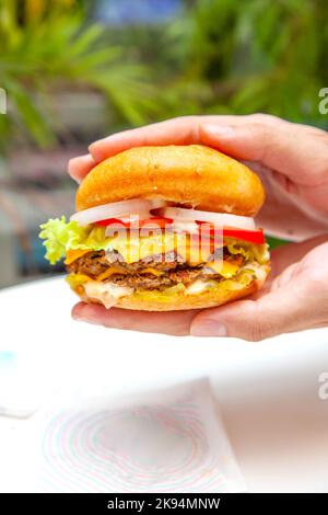 Hands about to lift a queso melted cheese burger with lush green background inside a cafe. Stock Photo