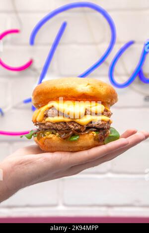 Hand palm holding a quesso melted cheese burger against a wall in cafe with neon sign. Stock Photo