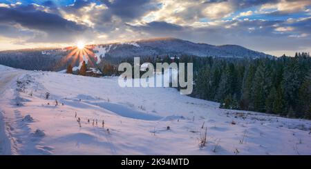 countryside landscape in mountains at sunset. beautiful scenery with coniferous forest on the snow covered hills in evening light. sun above the ridge Stock Photo