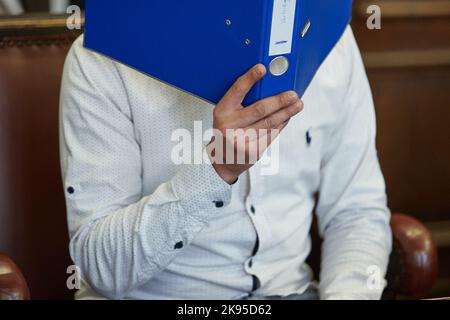 Hamburg, Germany. 26th Oct, 2022. The defendant sits in the courtroom before the trial begins, holding a blue file folder in front of his face. The 27-year-old is on trial for attempted manslaughter. The defendant is alleged to have fatally injured his partner with a knife in October 2021. He fled across adjacent rooftops and was arrested in Paris on November 11, 2021, on an international arrest warrant and transferred to Germany. Credit: Georg Wendt/dpa/Alamy Live News Stock Photo